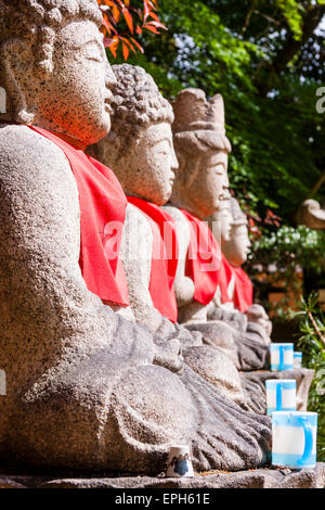 Vue rapprochée le long de quatre statues assises de Jozo bosatsu avec des bavoirs rouges en lumière, à l'extérieur au temple de Suma Dera à Suma, Japon. Banque D'Images