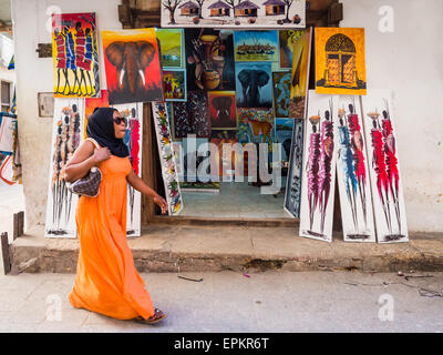 Femme marche devant un magasin vendant Tinga Tinga peintures colorées comme souvenirs aux touristes à Stone Town, Zanzibar. Banque D'Images