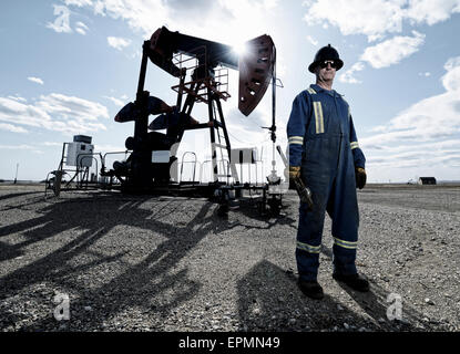 Un homme en bleu de travail et casque à une pompe à balancier en pleine terre à un site d'extraction d'huile. Banque D'Images