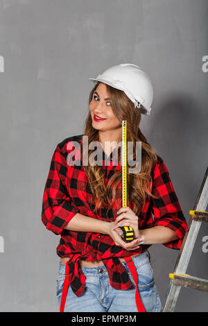 Jeune fille construction worker holding ruban à mesurer. Femme souriante. Fille Vêtue de jeans, chemise à carreaux et un casque. Banque D'Images