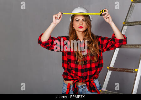 Jolie jeune fille builder tenant un ruban à mesurer. Fille Vêtue de jeans, une chemise à carreaux et un casque. Concept de la construction Banque D'Images