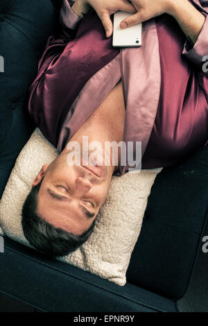 Homme étendu et se reposer sur un canapé à la maison. Il est en train de dormir avec sa tête sur un coussin et tenant un téléphone cellulaire dans les mains. Banque D'Images