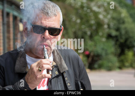 Homme d'âge moyen dans les lunettes de souffler sur un e-cigarette comme il se tient à l'extérieur dans l'avant-cour d'un bâtiment avec des arbres derrière h Banque D'Images