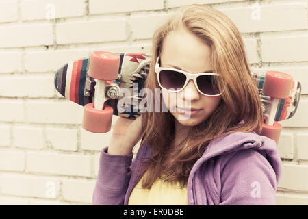 Adolescente blonde à lunettes détient près de skateboard urbain mur brique gris vintage, correction tonale, old style effet du filtre Banque D'Images