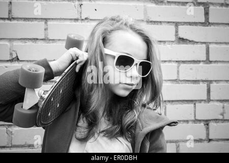 Adolescente blonde en jeans et lunettes détient plus de skateboard urban mur de briques, photo monochrome Banque D'Images
