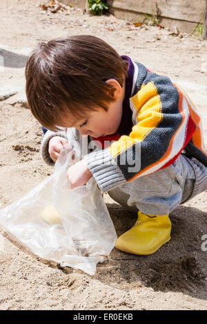 Jeune enfant, garçon, 3-4 ans, se brouiller à l'extérieur dans un parc en jouant avec un sac en plastique au sol. L'ouvrir et regarder à l'intérieur. Banque D'Images