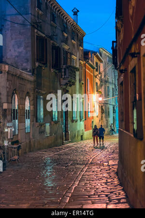 Soirée photo d'une rue pavée de Rovinj, Croatie. Un couple marche silhouetté contre le coloré, lampe-immeubles illuminés. Banque D'Images