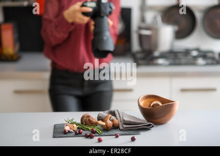 Une femme photographe prend un close-up de l'automne les fruits et légumes - champignons, l'ail, le romarin, et les canneberges. Sur le comptoir, un bol en bois. Une cuisinière à gaz et pot à l'arrière-plan. Banque D'Images