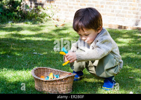 Enfant, garçon, 4-5 ans, aux rayons du soleil, se brouillant sur la pelouse dans le jardin tout en ramassant les patères et en les plaçant dans un panier en osier. Banque D'Images