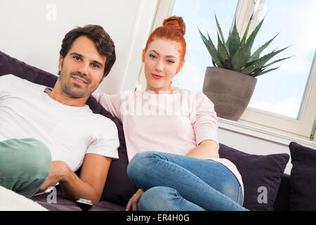 Jeune couple en appui sur le canapé dans le salon tandis que l'homme est de regarder la télévision et d'une femme est en train de lire un livre. Banque D'Images