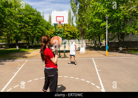 Jeune couple athlétique jouant au basket-ball ensemble - homme regardant femme prend tourné à partir du haut de la clé sur une cour, dans un parc verdoyant. Banque D'Images