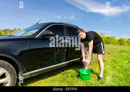 L'homme à la caméra et à l'essorage de l'Éponge savonneuse dans la benne verte pendant le lavage de véhicules de luxe noir en vert sur le terrain herbeux journée ensoleillée avec ciel bleu. Banque D'Images