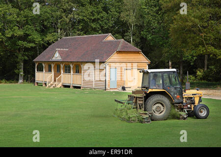 Un tracteur coupe de l'herbe en face d'un bois de construction traditionnelle construction pavillon cricket anglais dans le sud de l'Angleterre Banque D'Images