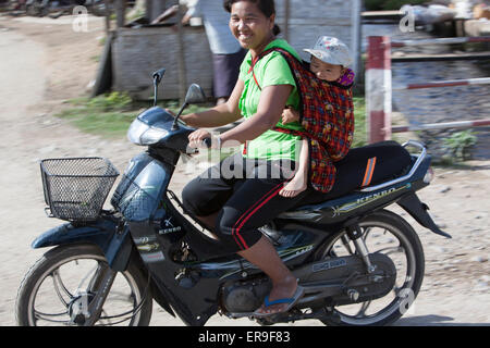 Une femme sur une moto sourit à la caméra comme elle chevauche passé. Son enfant est en bandoulière sur son dos dans un tissu de l'élingue. Banque D'Images