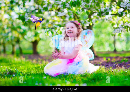 Enfant jouant avec un oiseau. Heureux rire petite fille en costume de fée avec des ailes de nourrir un perroquet dans un cerisier jardin Banque D'Images