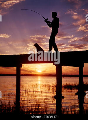 Années 1950 Années 1960 LA SILHOUETTE ANONYME GARÇON DE PÊCHE AVEC SON CHIEN SUR UN QUAI AU COUCHER ET AU LEVER DU SOLEIL Banque D'Images