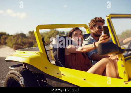 Heureux heureux couple enjoying road trip dans leur voiture. Homme conduisant la jeune femme en prenant une voiture de selfies son téléphone mobile. Banque D'Images