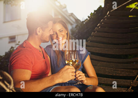 Belle jeune couple toasting with wine à l'extérieur. Ils sont assis sur un hamac de sourire et de boire du vin avec un soleil éclatant en b Banque D'Images