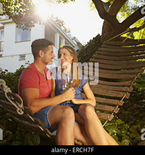 Young couple toasting with wine alors qu'il était assis sur un hamac en arrière-cour. Ils sont à la recherche à chaque autre smiling et boire Banque D'Images