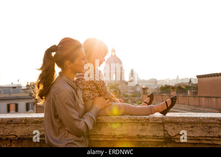 Au coucher du soleil, une brunette mère de profil, il est debout derrière elle, tenant sa fille blonde, comme elle est assise sur un rebord au-dessus Banque D'Images