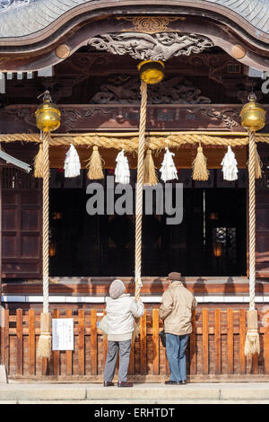 Japon, Matsumoto, Yohashira-jinja. La salle principale du temple, Haiden avec un couple âgé debout devant sonner le cordage de cloche pendant qu'ils prient. Banque D'Images