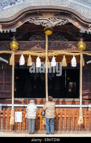 Japon, Matsumoto, Yohashira-jinja. La salle principale du temple, Haiden avec un couple âgé debout devant sonner le cordage de cloche pendant qu'ils prient. Banque D'Images