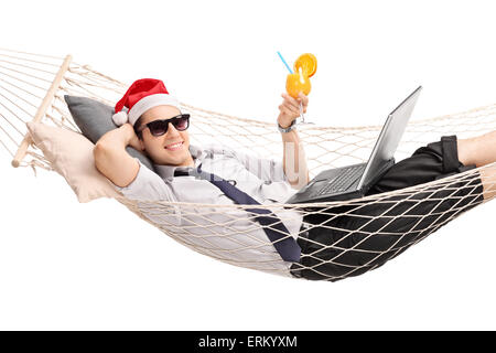 Young man with Santa hat couché dans un hamac avec un cocktail dans la main et un ordinateur portable sur ses genoux isolé sur fond blanc Banque D'Images