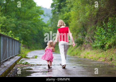 Mère et petit curly bébé fille marcher ensemble dans un parc sur une journée d'été humide après la pluie, vue de l'arrière Banque D'Images