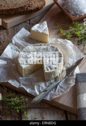 Un morceau de camembert sur une planche à découper, un couteau et des herbes. Banque D'Images