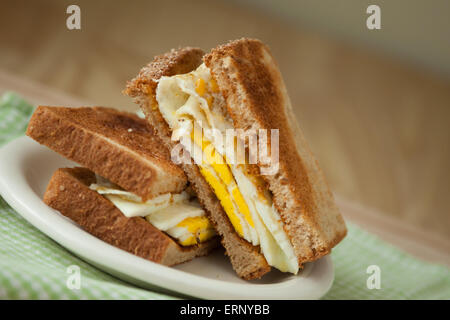 Un œuf frit sandwich sur pain de grains entiers est assise sur une assiette blanche au sommet d'une serviette à carreaux blancs et verts et une planche à découper en bois Banque D'Images