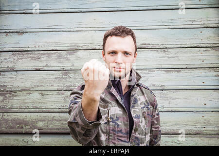 Young Caucasian man in camouflage montrant son gros poing. Portrait en extérieur sur green rural mur en bois Banque D'Images