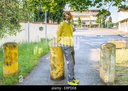 Bel homme d'âge moyen avec des cheveux poivre sel, moyenne, cheveux habillés en vêtements décontractés avec chandail jaune, un pantalon bleu et jaune vert baskets dans l'extérieur : il montre un regard rassurant tout en coin sur bollard Banque D'Images