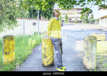 Bel homme d'âge moyen avec des cheveux poivre sel, moyenne, cheveux habillés en vêtements décontractés avec chandail jaune, un pantalon bleu et jaune vert baskets dans l'extérieur : il montre un regard rassurant tout en coin sur bollard Banque D'Images