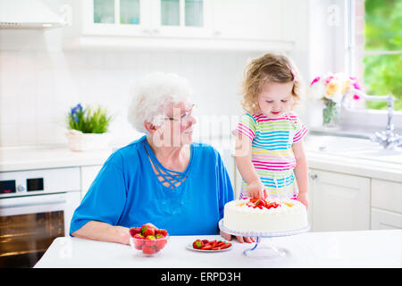 Happy senior dame, grand-mère aimante, la confection d'un gâteau aux fraises fait maison avec sa petite-fille dans une cuisine moderne blanc Banque D'Images