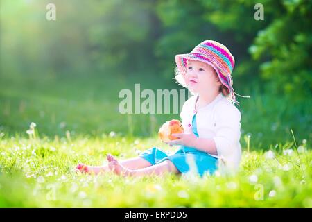 Heureux belle petite fille portant une robe bleue et chapeau de paille colorée big apple comme manger frais collation santé assis sur la pelouse Banque D'Images