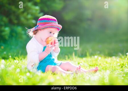 Heureux belle petite fille portant une robe bleue et chapeau de paille colorée big apple comme manger frais collation santé assis sur la pelouse Banque D'Images