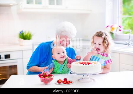 Happy senior dame, grand-mère aimante, la confection d'un gâteau aux fraises fait maison avec deux enfants dans une cuisine moderne blanc avec fenêtre Banque D'Images