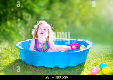 Happy cute girl avec des cheveux bouclés portant une robe colorée rose jouant dans un bac à sable avec des jouets en plastique billes dans jardin Banque D'Images