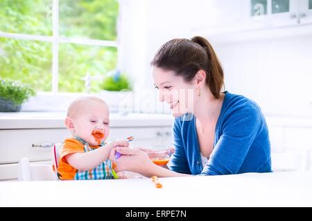 Young attractive mère allaiter son bébé mignon fils, lui donnant son premier aliment solide, saine de pure végétale carotte Banque D'Images