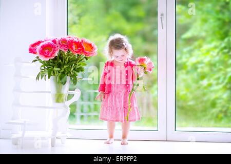 Heureux mignon bébé fille avec des cheveux bouclés portant une robe rose de jouer avec un tas de belles grosses pivoines dans un vase Banque D'Images