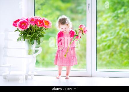 Heureux mignon bébé fille avec des cheveux bouclés portant une robe rose de jouer avec un tas de belles grosses pivoines dans un vase Banque D'Images