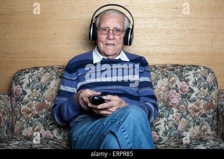 Parution du modèle. Senior man sitting on sofa portant des écouteurs à regarder la télévision. Banque D'Images