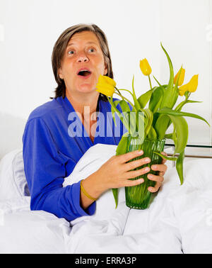 Femme à la cinquantaine assis au lit et tenant un vase de fleurs Banque D'Images