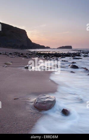 Mer calme, clapotis des vagues, le sable, côte rocheuse, silhouetté falaises & Saltwick Nab sous Red sky at sunset - Saltwick pittoresque baie tranquille, Yorkshire, UK. Banque D'Images