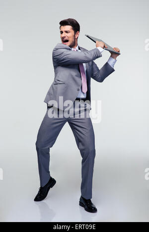 Portrait de angry businessman essaie de détruire son ordinateur portable sur fond gris Banque D'Images
