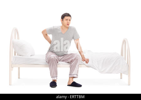 Jeune homme ayant un mal de dos assis sur un lit dans son pyjama isolé sur fond blanc Banque D'Images