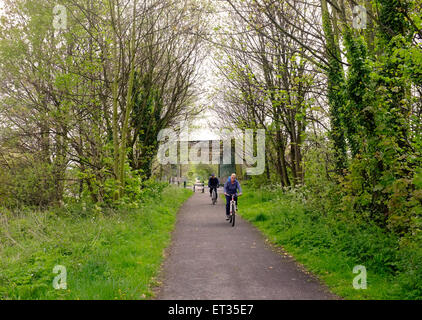 Deux cyclistes équitation le long d'un chemin avec des arbres et l'herbe. Pont et porte dans la distance. Banque D'Images