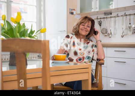 Happy senior woman having une tasse de thé et talking on cell phone in kitchen, Munich, Bavière, Allemagne Banque D'Images