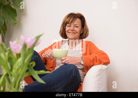 Happy senior woman sitting on sofa et d'avoir une tasse de thé à la maison, Munich, Bavière, Allemagne Banque D'Images