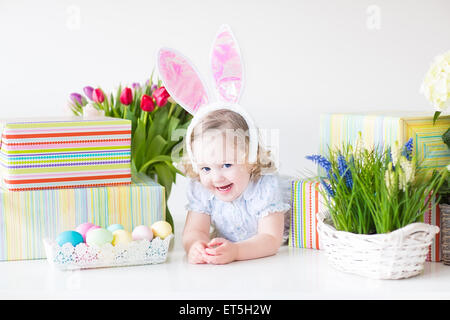 Happy laughing toddler girl wearing a blue dress et oreilles de lapin de Pâques avec des oeufs colorés, présente et fleurs de printemps Banque D'Images